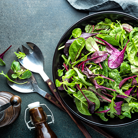 A mix of italian green and red lettuces, served in a rustic bowl. To the left are salad tongs and other wooden-handled utensils.
